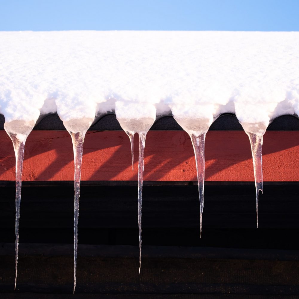 Long icicles hanging from the black and red roof of a cabin in the snow against a blue sky copy spac